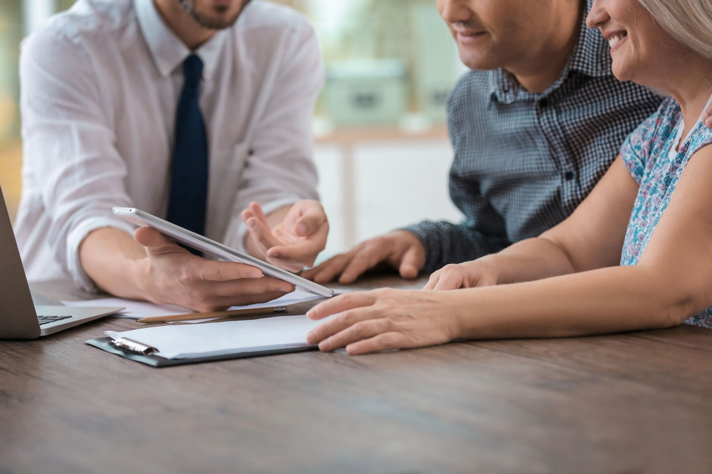 Insurance agent showing a tablet to a smiling couple while discussing documents at a wooden table.