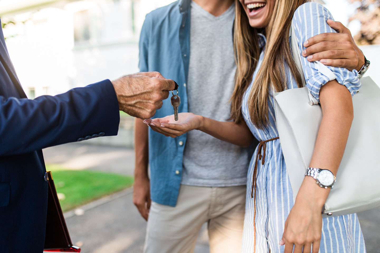 Smiling couple receiving house keys from a person in a blue blazer outdoors.
